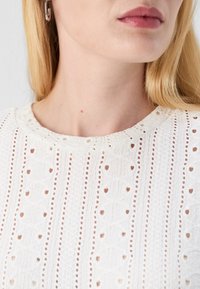 White textured top with a round neckline, featuring intricate embroidered patterns and small cutouts throughout the fabric.