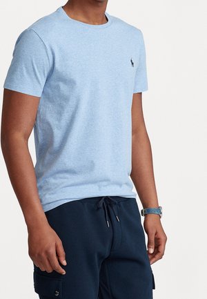 Mężczyzna ubrany w jasnoniebieski t-shirt z krótkim rękawem i okrągłym dekoltem, z małym czarnym logo na piersi, oraz granatowe spodnie na sznurku z bocznymi kieszeniami.