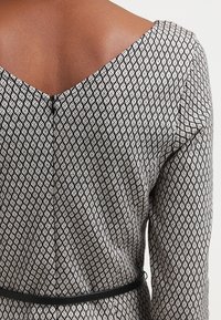 Robe à motifs noir et blanc avec un dos décolleté en V profond, un motif en forme de losange, des manches longues et une ceinture noire à la taille.