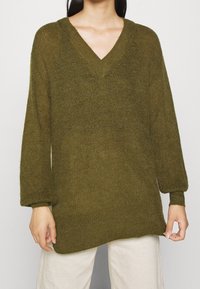 Personne portant un pull en maille poilue vert olive à encolure en V avec des manches longues et un pantalon de couleur crème sur un fond neutre.