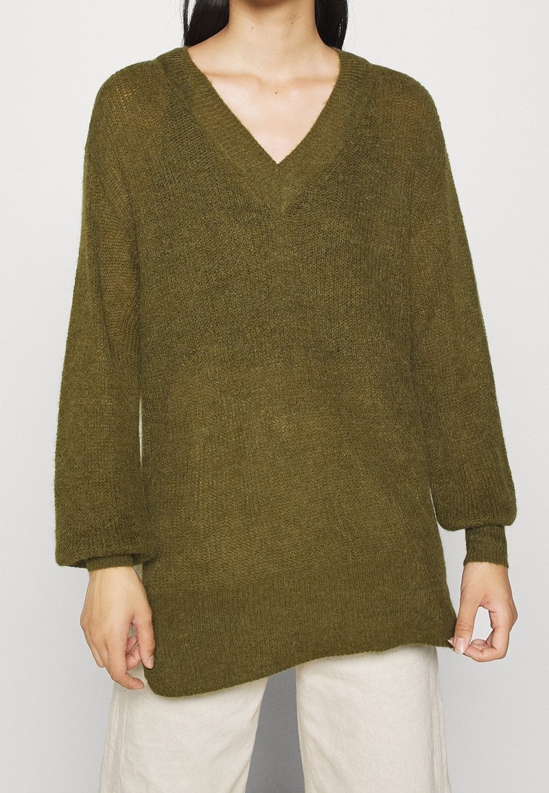 Personne portant un pull en maille poilue vert olive à encolure en V avec des manches longues et un pantalon de couleur crème sur un fond neutre.