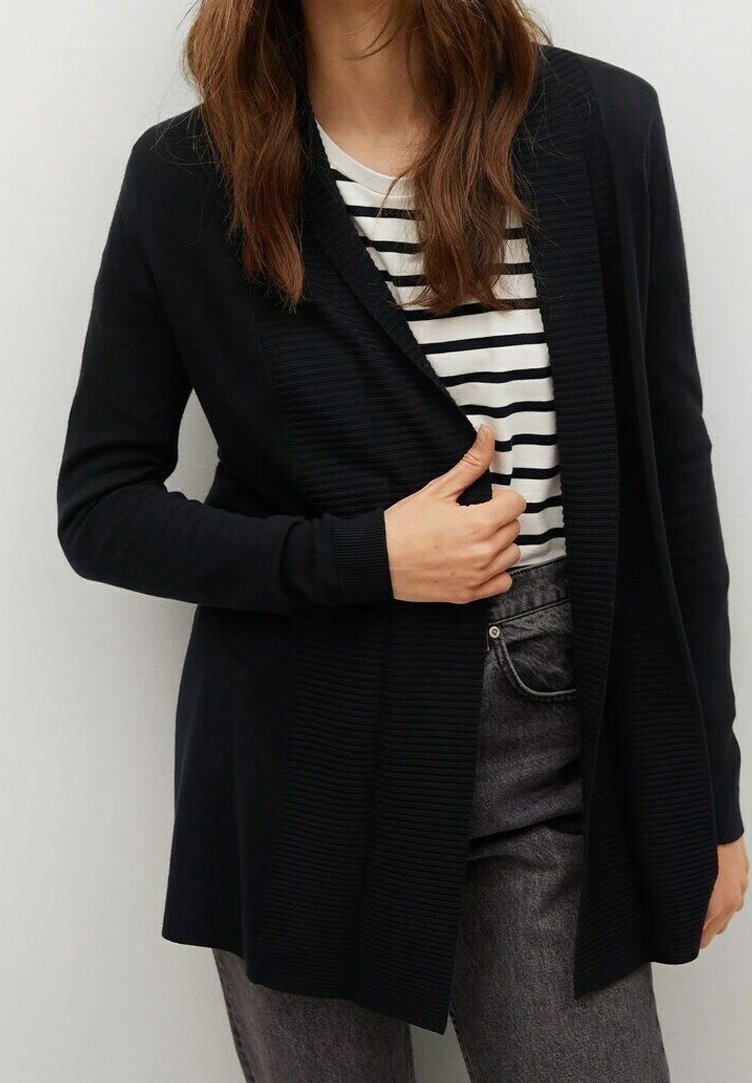 Femme portant un cardigan noir sur une chemise rayée blanche et noire et un jean gris, tenant le cardigan fermé au niveau de la poitrine.