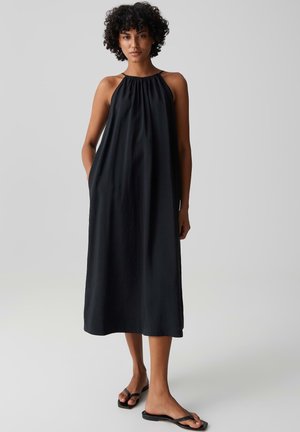 Femme aux cheveux bouclés portant une robe midi noire sans manches et ample, ainsi que des sandales slides noires, debout devant un fond clair uni.