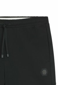 Pantaloni jogger neri con vita regolabile, caratterizzati da un pannello laterale testurizzato nero e un patch logo esagonale in una tonalità attenuata.