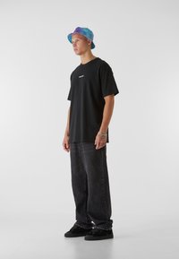T-shirt noir avec logo, associé à un jean gris foncé ample et des baskets noires. Le mannequin porte un chapeau de seau multicolore.