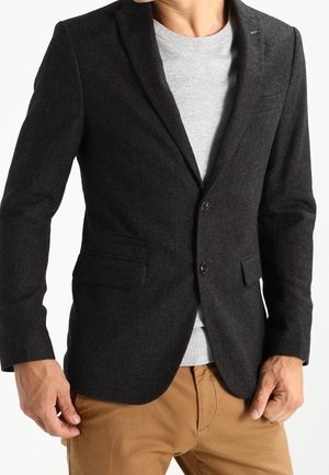 Mężczyzna w ciemnoszarej marynarce na jasnoszarym swetrze i beżowych spodniach, stojący na białym tle.