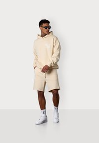Haut à capuche beige avec un logo sur le devant, associé à des shorts beiges. Le mannequin porte des lunettes de soleil, des baskets blanches et des chaussettes blanches à motifs. Texture lisse.