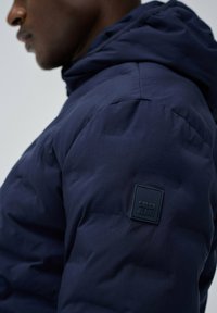 Tamno plava puf jakna s teksturiranim završetkom, koja sadrži modernu kapuljaču i gumirani logo "Salsa Jeans" na ramenu.