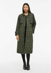 Kvinne i lang, mørkegrønn quiltet frakk med knapper, svarte leggings og svarte ankelstøvler, står mot en hvit bakgrunn.