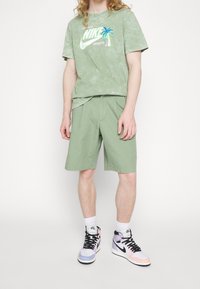 Osoba w zielonej koszulce Nike z grafiką palmy, dopasowanych zielonych szortach, białych skarpetach Nike i pastelowych sneakersach Nike.