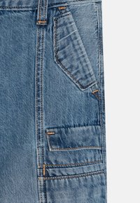 La tela de mezclilla azul claro presenta un diseño de bolsillo distintivo con costuras naranjas en contraste. La textura parece ligeramente desgastada con un desvanecimiento sutil.