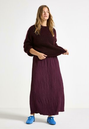 Burgunderfärgad, strukturerad stickad tröja med överdimensionerade ärmar, parat med en lång, plisserad lila kjol. Klara blå sneakers fulländar outfiten.