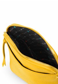 Žlutá kožená taštička se zlatým zipem, otevřená, odhalující černou látkovou podšívku s vzorem z vícebarevných teček a nápisem "BE VUCH".