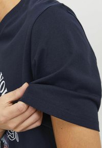 T-shirt en coton navy à manches courtes, mettant en avant le détail des poignets. Le tissu semble lisse avec une coupe ajustée, présentant des accents de design minimalistes.