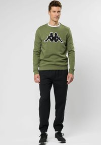Olijfgroene sweatshirt met zwart Kappa-logo, ronde halslijn en geribde manchetten, gecombineerd met zwarte taps toelopende joggers en zwarte sportschoenen.