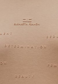 Μπεζ ύφασμα με ανάγλυφα, ανοιχτόχρωμα γράμματα που περιλαμβάνουν τα "Elisabetta Franchi", "Determination" και διάφορες άλλες λέξεις. Λείο, ελαστικό ύφασμα.