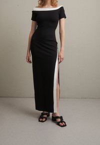 Czarna, dopasowana maxi sukienka z białym dekoltem off-shoulder i rozcięciem z boku. Miękki materiał i elegancki krój. Zestawiona z czarnymi sandałami na ramiączkach.