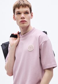 Ružové bavlnené tričko s okrúhlym krémovým logom na hrudi. Modelka drží cez jedno rameno čierny popruh tašky.