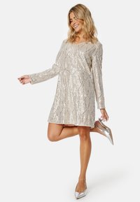 Šaty se sequinovým zdobením, v výstřihu do V, s dlouhými rukávy a volným střihem. Mají stříbrný a béžový vzor. S outfitem ladí stříbrné lodičky se špičatou špičkou.
