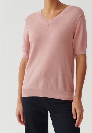 Femme portant un haut en maille rose clair à manches courtes avec un col en V, associé à un pantalon foncé, debout devant un fond gris uni.