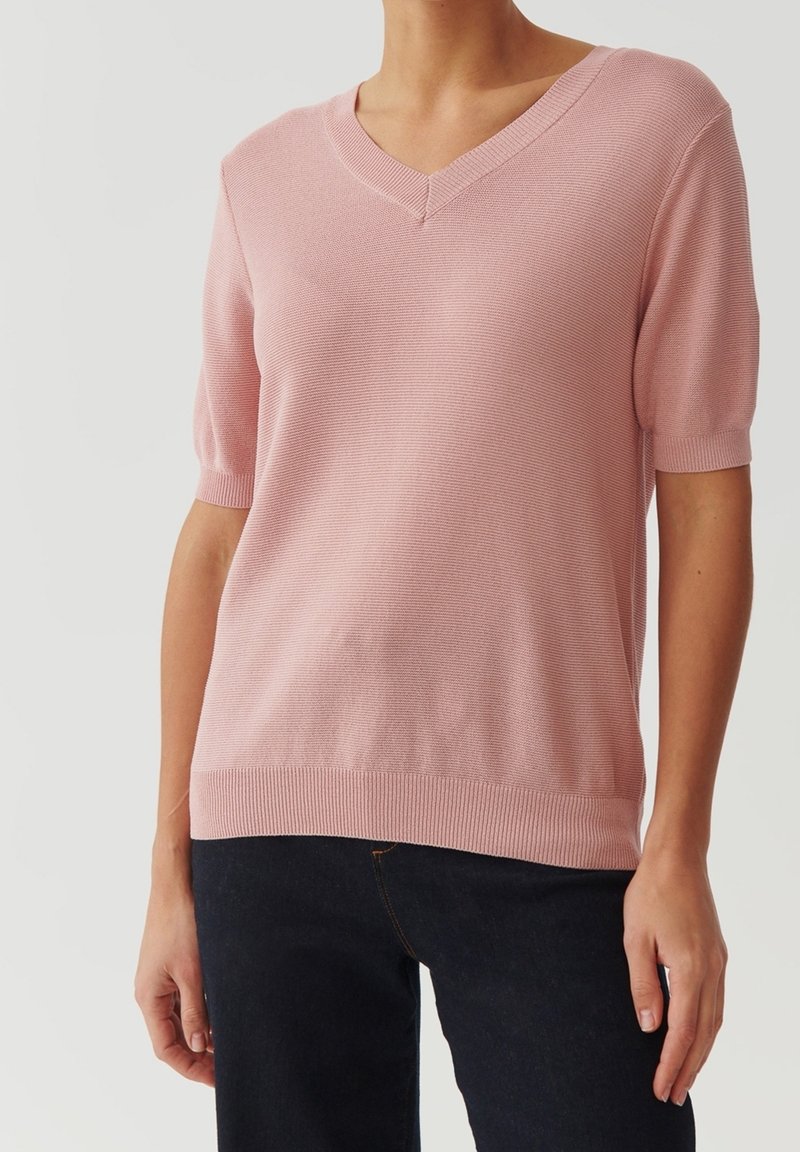 Femme portant un haut en maille rose clair à manches courtes avec un col en V, associé à un pantalon foncé, debout devant un fond gris uni.