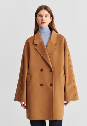 Jeune femme aux cheveux bruns raides porte un manteau ample croisé couleur camel par-dessus un col roulé bleu clair.