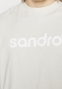Gros plan sur une personne portant un t-shirt blanc avec des lettres blanches en relief formant "sandro" sur la poitrine.