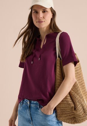 Frau mit weißer Kappe, lila Kurzarmtop, blauen Jeans und trägt eine große, geflochtene beige Umhängetasche auf der Schulter.