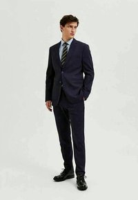 Completo blu con un motivo testurizzato, caratterizzato da una giacca aderente e pantaloni sartoriali. Abbinato a una camicia azzurro chiaro e una cravatta a righe. Scarpe nere.