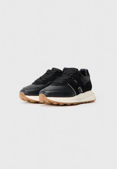 Adidași negri cu un finisaj din piele, având un accent din suede, vârf rotund, guler căptușit și o talpă din cauciuc de culoare deschisă cu textură antiderapantă.