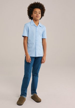 Cămașă bleu, cu mânecă scurtă și nasturi, într-o culoare solidă, asortată cu blugi din denim albastri și pantofi slip-on maro, stând în fața unui fundal neutru.