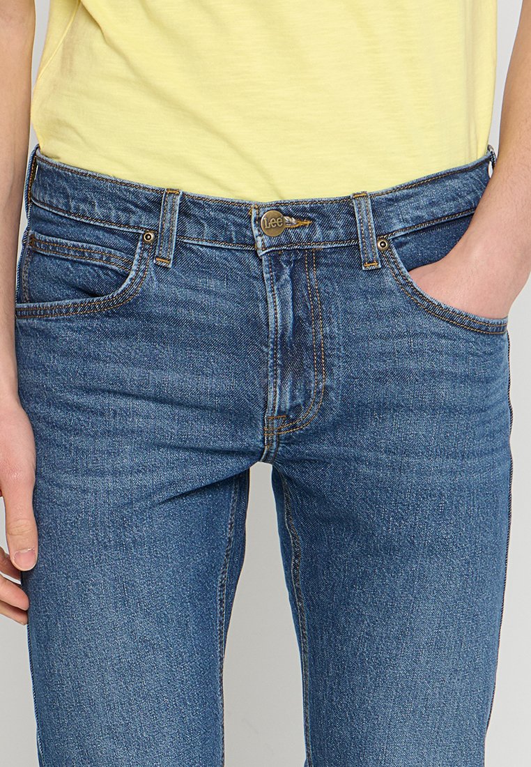 Osoba w niebieskich dżinsach z guzikami i żółtej koszuli, z prawą ręką w kieszeni, a lewą ręką spoczywającą obok.