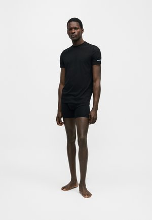 Modelo masculino de pie, descalzo, vestido con una camiseta negra ajustada y bóxers negros frente a un fondo claro y liso.