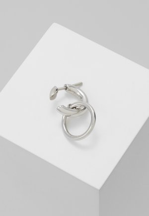 Stříbrná kovová kruhová náušnice s menším kruhem propojeným s hlavním kruhovým tvarem, umístěná na bílém povrchu.