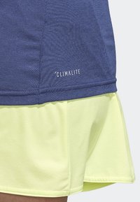 Blå atletisk topp med 'Climalite'-etikett, i kombination med en ljusgul lagerkjol. Materialen verkar vara släta med en blandning av texturer.