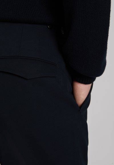 Dark navy pants with a smooth fabric, featuring a back pocket detail and two black buttons at the waist, paired with a knitted top.