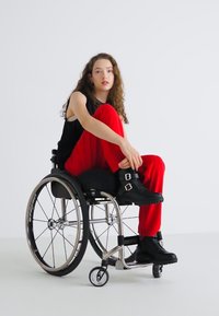 Black sleeveless top, red wide-leg trousers, and black ankle boots. Model in a silver wheelchair with large wheels on a plain white background.