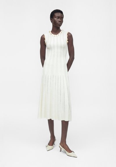 Femme mince à la peau foncée, debout les mains dans le dos, portant une robe midi blanche en maille sans manches avec une bordure fleurie et des chaussures blanches à talons bas.