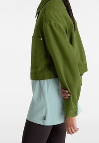 Giacca di jeans verde oliva cropped con colletto a punta, tasca laterale e polsini con bottone, indossata sopra una camicia azzurro chiaro e leggings scuri.