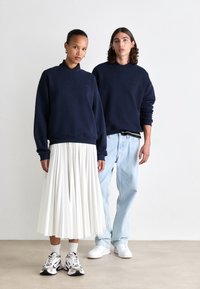 Dunkelblaue Sweatshirts mit rundem Halsausschnitt, kombiniert mit einem weißen, plissierten Rock und hellblauen Jeans. Beide tragen sportliche Sneakers.