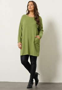 Groene oversized trui met lange mouwen en voorzakken, gecombineerd met zwarte leggings en puntige enkellaarsjes. Glad textiel, ontspannen pasvorm.