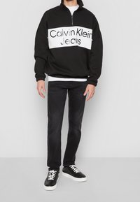 Black zip-up sweatshirt with white horizontal stripe featuring "Calvin Klein Jeans" text, paired with dark jeans and black sneakers.