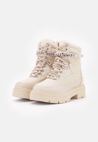 Bottes d"extérieur beiges avec une finition texturée, dotées de quincaillerie argentée, de détails en dentelle et d"une semelle épaisse et robuste. Accents élégants en chaîne sur le dessus.