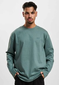 Green long-sleeve t-shirt in a soft, textured fabric. Features a crew neck and a subtle embroidered logo on the chest. Relaxed fit.