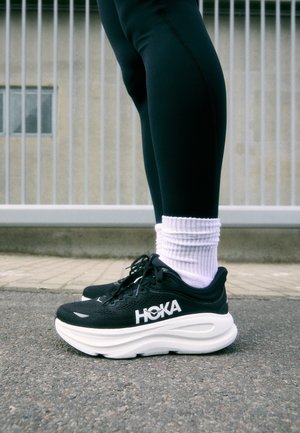 Person trägt schwarze HOKA Laufschuhe mit dicken weißen Sohlen, weiße Socken und schwarze Leggings und steht auf einer gepflasterten Fläche in der Nähe eines Metallzauns.