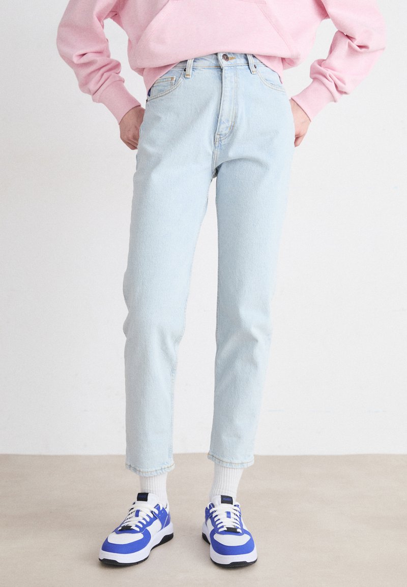 Jeans de tiro alto en azul claro con un ajuste estrecho, combinados con zapatillas blancas y azules. Los jeans presentan una textura de denim suave y descolorida.