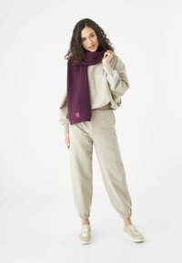 Vrouw met krullend haar, gekleed in beige sweatpants, een bijpassende sweatshirt, beige sneakers en een lange paarse sjaal, staand op een witte achtergrond.