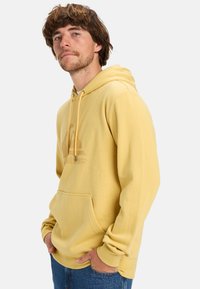 Sudadera amarilla con bolsillo frontal, capucha con cordón y un logo sutil impreso en un tono más claro. Puños acanalados y corte holgado.