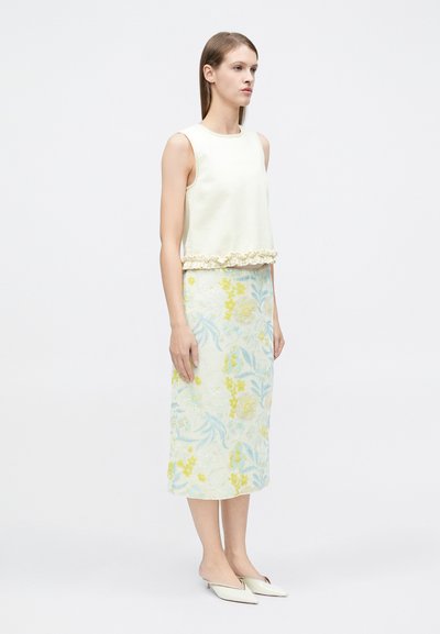 Femme debout portant un haut sans manches crème avec bordure à volants, une jupe midi fleurie en bleu pastel et jaune, et des mules pointues crème.