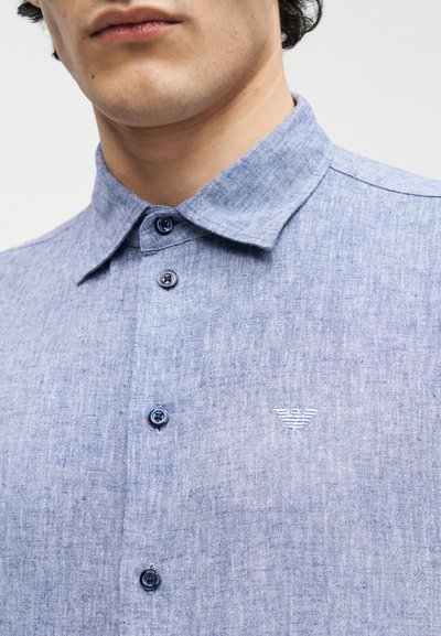 Άνδρας που φοράει ανοιχτό μπλε πουκάμισο με κουμπιά και ένα μικρό λευκό κεντημένο λογότυπο στο στήθος, ορατό από το λαιμό μέχρι τη μέση.